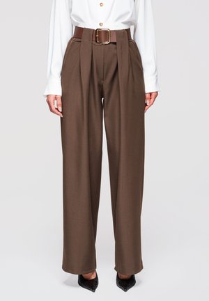 Pantalon marron à taille haute et jambes larges avec plis à l'avant, associé à une ceinture marron et des chaussures noires à bouts pointus, porté avec une chemise blanche boutonnée.