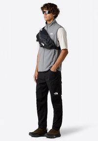 Šedá fleecová vesta s černou ledvinkou, černé cargo kalhoty a hnědé boty. Model nosí sluneční brýle a představuje tak ležérní venkovní outfit.