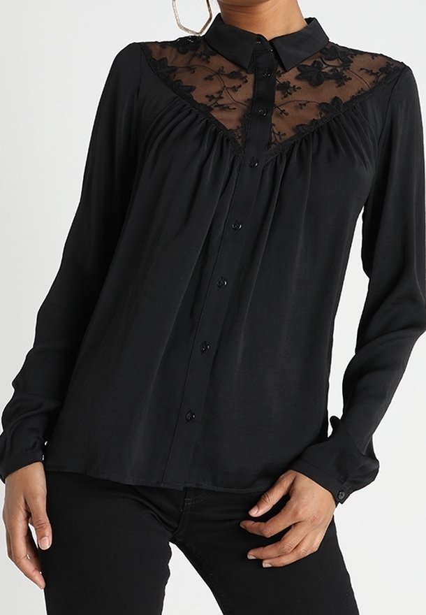 Blouse noire à manches longues avec boutonnage, empiècement en dentelle florale et col, portée avec un pantalon noir, et de grandes boucles d'oreilles visibles.