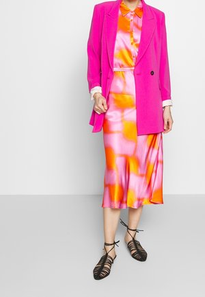 Heller pinker Blazer über einem bunten, satinartigen Kleid mit orange und pinken abstrakten Mustern; schwarze, sandalenartige Schuhe mit Riemchen vervollständigen das Outfit.