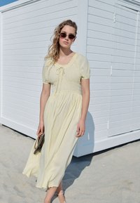 Gul midiklänning med puffade ärmar, en smockad överdel och en rundad ringning. Texturerat gingham-mönster, bärs med runda solglasögon och en väska.