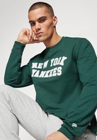 Grønn hettegenser med "NEW YORK YANKEES" trykket i hvitt, sammen med grå joggebukser. Har mannskapsnakke og lange ermer.