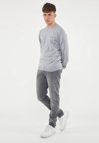 Camicia grigia a maniche lunghe con scollatura rotonda e piccolo logo, abbinata a jeans grigi e sneakers bianche. Design semplice, texture morbida.