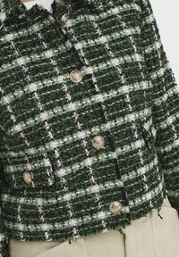 Veste texturée à carreaux verts et blancs avec des boutons décoratifs dorés, portée sur un pantalon beige, montrant le torse avant et les détails des poches.