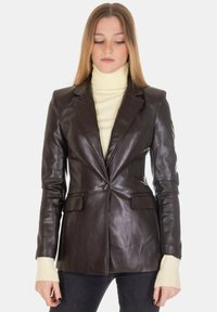 Blazer in pelle marrone con taglio su misura, revers a lancia, chiusura con un bottone e due tasche frontali. Indossato sopra un dolcevita color crema.