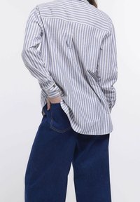 Pasiasta koszula zapinana na guziki w kolorze niebieskim i białym, wykonana z lekkiego materiału, z luźnym krojem i zaokrąglonym dołem, połączona z ciemnymi dżinsami.