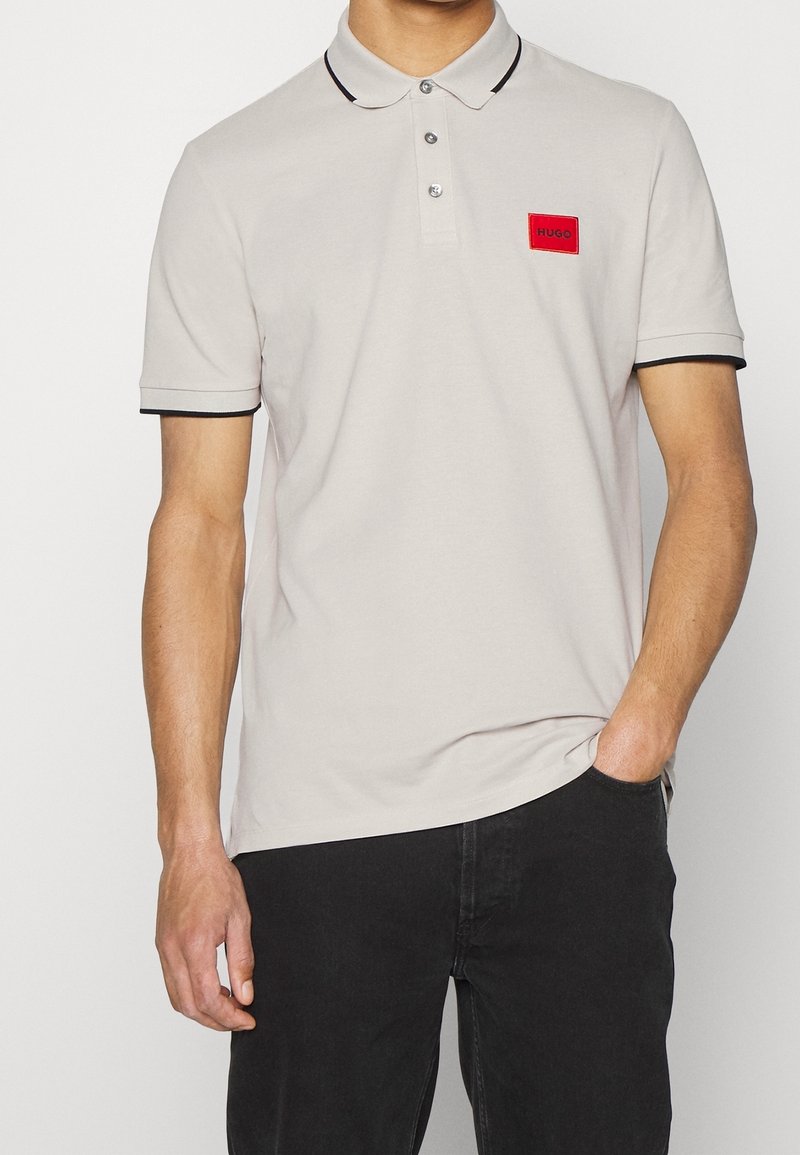 Homme portant un polo gris clair avec un bord noir au col et aux manches, écusson logo rouge "Hugo", et pantalon noir avec une main dans la poche.