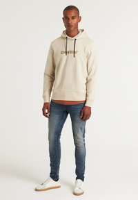 Beige kapucnis pulóver "CHASIN'" logóval, fekete húzózsinórokkal, kék skinny farmernadrággal és fehér sportcipővel, amelynek talpa bézs színű.