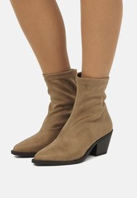 Piernas luciendo botas de ante beige de media caña con punteras puntiagudas y tacones negros gruesos sobre un fondo blanco.