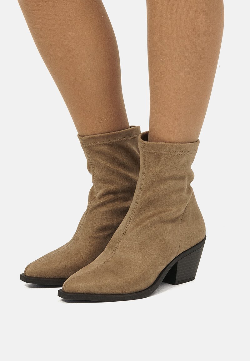 Piernas luciendo botas de ante beige de media caña con punteras puntiagudas y tacones negros gruesos sobre un fondo blanco.