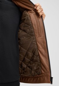 Bruine leren jas met een gevoerde bruine voering, voorzien van een ritssluiting en een zijzak aan de binnenkant, met een gestructureerd buitenoppervlak.