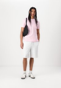 T-shirt rose clair avec une poche sur le devant, short gris et sac banane noir. Porté avec des baskets blanches, affichant une tenue décontractée.