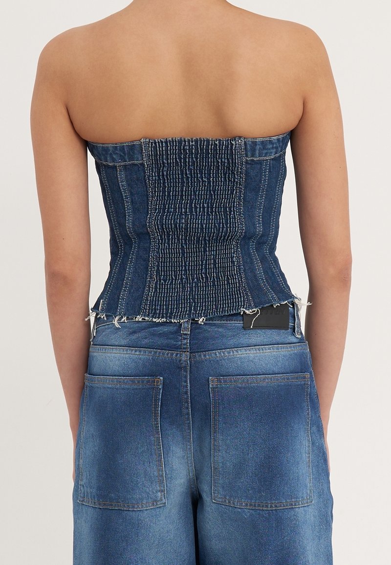 Riflový top bez ramínek se stahovaným zadním dílem, s konstrukčním designem a nedokončeným lemem. Nosený s riflemi s vysokým pasem.