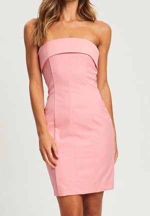 Femme portant une robe rose ajustée, sans bretelles, longueur genoux, avec un décolleté rabattable, debout devant un fond clair uni.