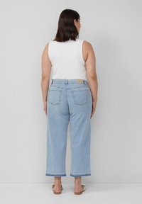 Lichtblauwe wijde jeans met hoge taille, achterzakken en een rafelige zoom. Draagt een witte mouwloze top en sandalen.
