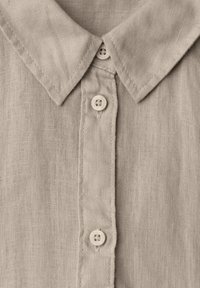 Gros plan du col d'une chemise en lin beige avec trois boutons cousus sur la patte de boutonnage, montrant la texture du tissu et les détails des coutures.