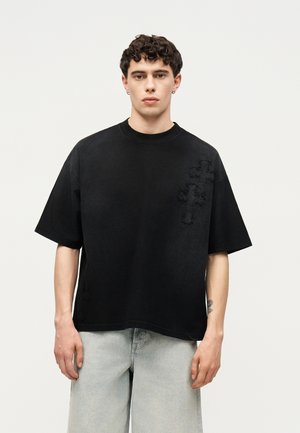 Jeune homme aux cheveux courts et bouclés portant un t-shirt noir oversized avec un motif croix texturé et un jean gris clair, debout sur un fond blanc.