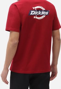 Camiseta de algodón roja de cuello redondo con un diseño de logotipo en blanco y negro de "Dickies" en la parte posterior. Mangas cortas y corte regular, textura suave.