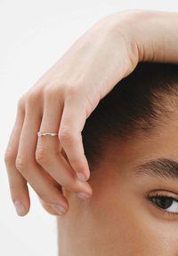 Anillo de plata con un acento de diamante, llevado en una mano que descansa cerca de la cara. La piel muestra una textura natural y suave. Fondo neutro.