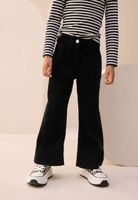 Schwarze Cord-Flared-Hose mit hoher Taille, kombiniert mit einem gestreiften Langarmshirt und weißen Sneakern mit Leopardenmuster.