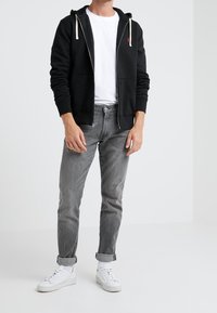 Sudadera negra con cremallera y capucha, con cordones blancos; combinada con jeans grises y zapatillas blancas. Presenta un diseño sencillo y un corte casual.