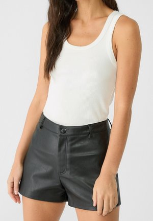 Kvinde iført hvid ærmeløs ribbet top og sorte højtaljede lædershorts med knapper foran og bæltestropper.