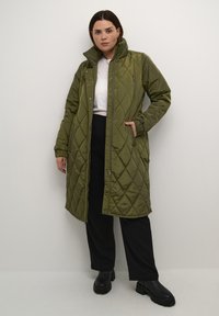 Palton lung matlasat de culoare verde măslinie, cu guler înalt, închidere cu capse și două buzunare laterale. Asortat cu un tricou alb și pantaloni negri.