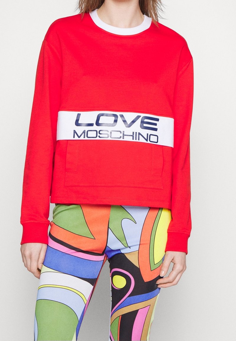 Femme portant un sweat-shirt rouge à manches longues avec le texte "LOVE MOSCHINO" et des leggings à motifs abstraits colorés.