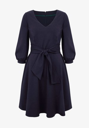 Marineblauwe jurk met V-hals, driekwart pofmouwen, getailleerde taille en bijpassende riem die vooraan in een strik is gebonden.