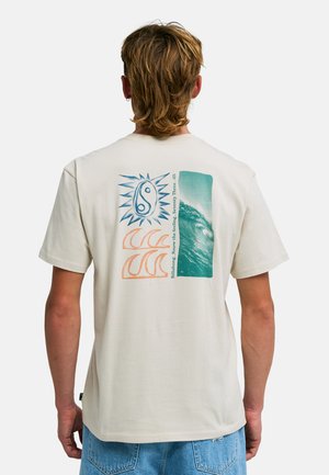 Uomo che indossa una t-shirt color crema con disegni blu di sole e onde e testo, abbinata a pantaloncini di jeans azzurri, di spalle.