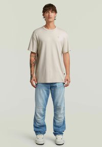 Beige t-shirt med kort ärm och rund hals, kombinerad med ljusblå slitna jeans och vita sneakers. Har en diskret logotyp på t-shirten.