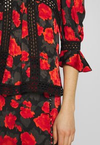 Schwarze Bluse mit floralem Muster und roten Blumen, mit einem mehrlagigen Design, Spitzenbesatz und ausladenden Ärmeln; kombiniert mit einem passenden Rock mit Knöpfen.