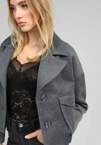 Manteau en mélange de laine gris avec de grands revers, deux boutons et une poche poitrine, porté par-dessus un haut en dentelle noir et un jean noir taille haute.