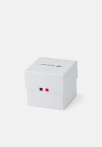 Caja cuadrada blanca con tapa, que presenta el logo de Lacoste y tres franjas de colores (azul, blanco, rojo) en el frente. Textura suave.