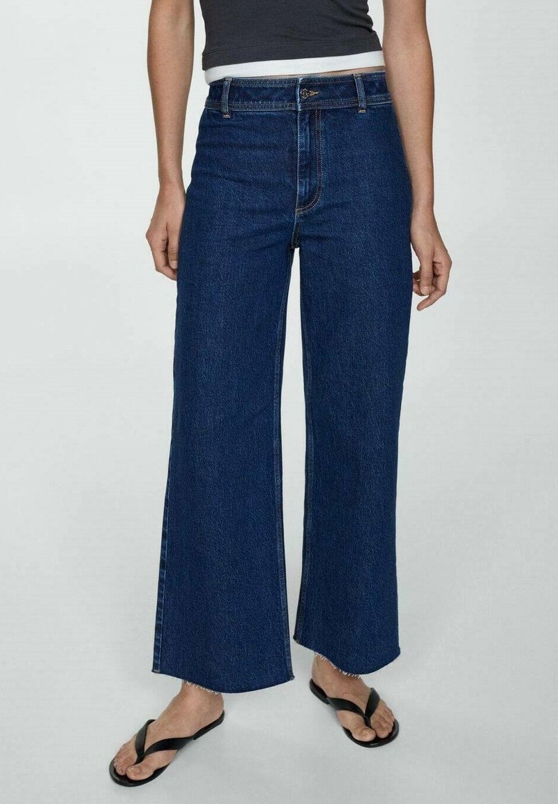 Jean à jambes larges en denim foncé, avec une taille haute, un design classique à cinq poches et des ourlets effilochés. Associé à des tongs noires.