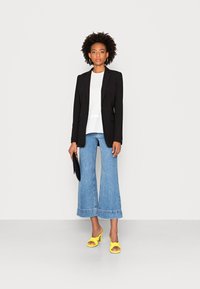 Blazer preto bem cortado, blusa branca com bainha costurada, calças de ganga largas em azul, e sandálias amarelas de salto alto. Os acessórios incluem uma clutch preta.