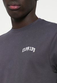 T-shirt cinza de manga curta com um logotipo em texto branco curvado no peito. O tecido parece suave, com um ajuste regular e gola redonda clássica.