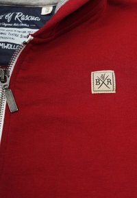 Red zip-up sweatshirt with a smooth texture, gray collar, and a rectangular, beige brand patch featuring logo and crossed symbols.