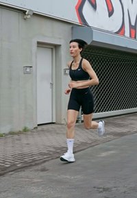 Svart sportbh og shorts, hvite sokker og joggesko. Personen jogger på en asfaltoverflate nær en grå vegg med graffiti.
