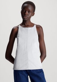 Hvit ermeløs topp med teksturert overflate, med rund halslinning og smale stropper, kombinert med blå jeans.