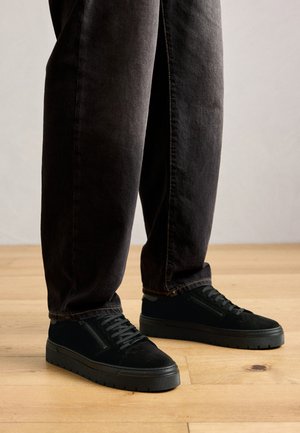 Personne portant des baskets noires avec des lacets et des zippers sur le côté, associées à un pantalon sombre ample, debout sur un sol en bois.