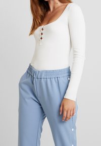 Femme portant un haut blanc à manches longues côtelé avec des boutons sur le devant et un pantalon taille haute bleu clair avec des boutons-pression sur les côtés.