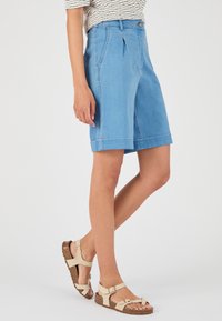 Pantalones cortos de mezclilla azul claro tipo bermuda con pliegues frontales, bolsillos laterales y puños enrollados, combinados con sandalias de tiras beige.