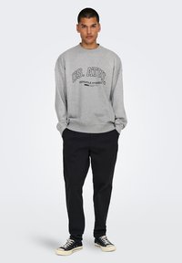 Gray sweatshirt with "OS. ATHL" print, round neckline, and long sleeves paired with black trousers and black sneakers with white soles.
