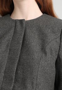 Chaqueta de mezcla de lana gris oscuro con cuello alto, pliegues sutiles y hombros estructurados. La textura parece suave con un corte ajustado.