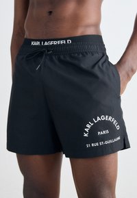Juodi maudymosi šortai su elastingu juosmeniu ir raišteliu, balta logotipo ir teksto akcentais, lygaus audinio, iki vidurinės šlaunies ilgio, be kišenių.