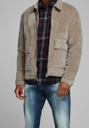 Mand iført beige manchesteroverdel over rød og navy plaid skjorte med falmede blå jeans, stående mod grå baggrund.