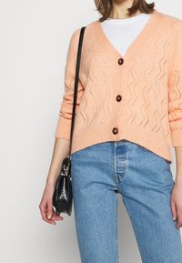 Melounově růžový pletený cardigan s výstřihem do V, vlnitým vzorem a třemi hnědými knoflíky. V kombinaci s světle modrými džíny a malou černou kabelkou.