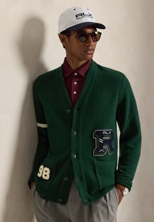 Jeune homme portant un cardigan vert boutonné avec un écusson "R", polo bordeaux, pantalon gris, casquette blanche RLX, lunettes de soleil et montre-bracelet.