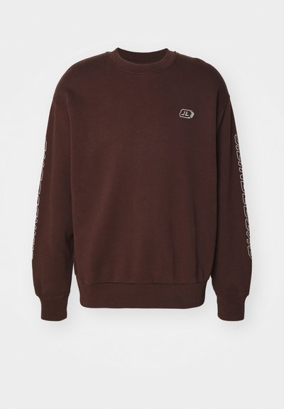 Καφέ φούτερ με στρογγυλή λαιμόκοψη, μακριά μανίκια και ριμπ μανσέτες. Έχει ένα μικρό λογότυπο στο στήθος και κείμενο κατά μήκος του μανικιού.